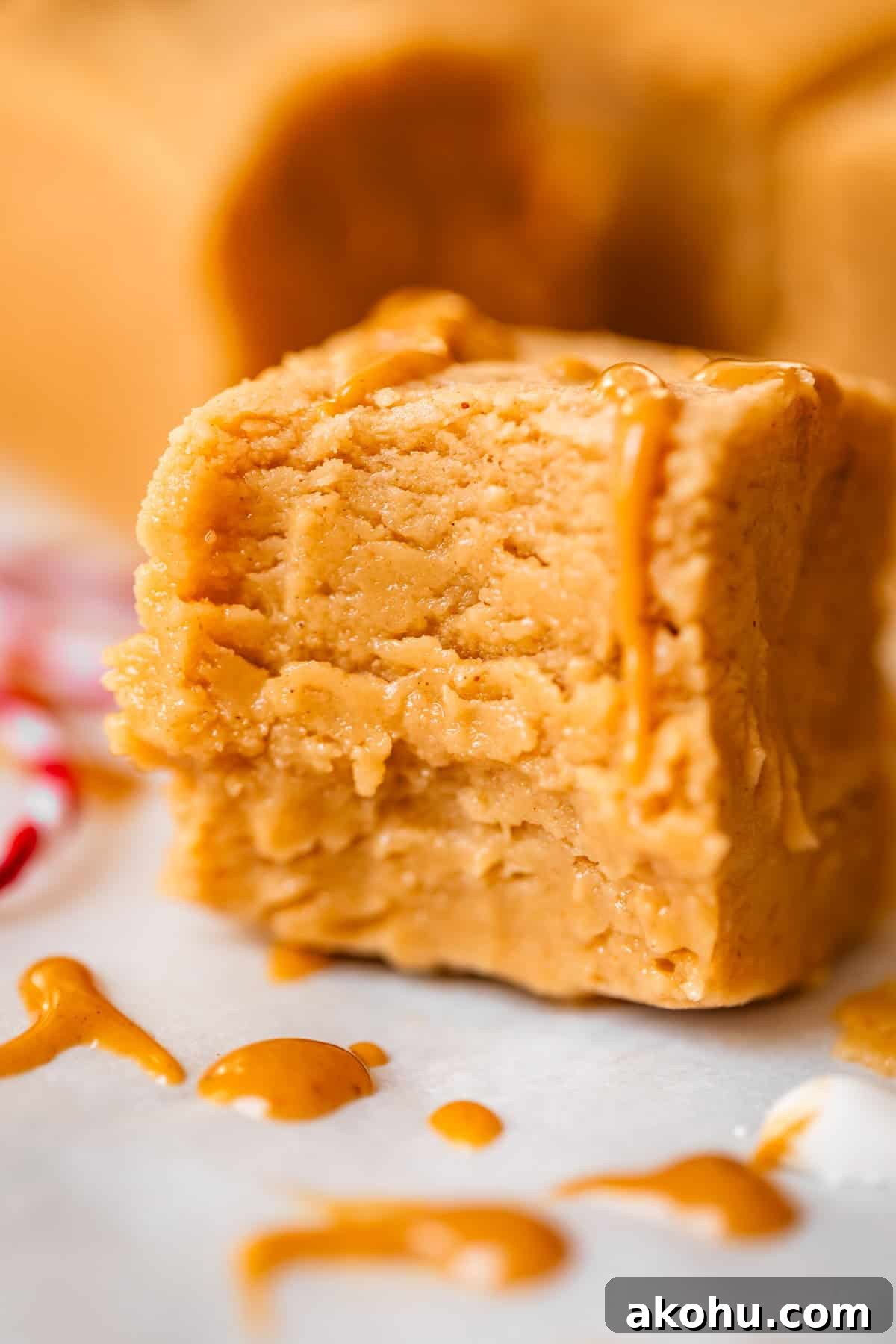 A close-up of a thick, creamy square of peanut butter fudge with a drizzle of melted peanut butter on top, sitting on parchment paper with extra drips around it.