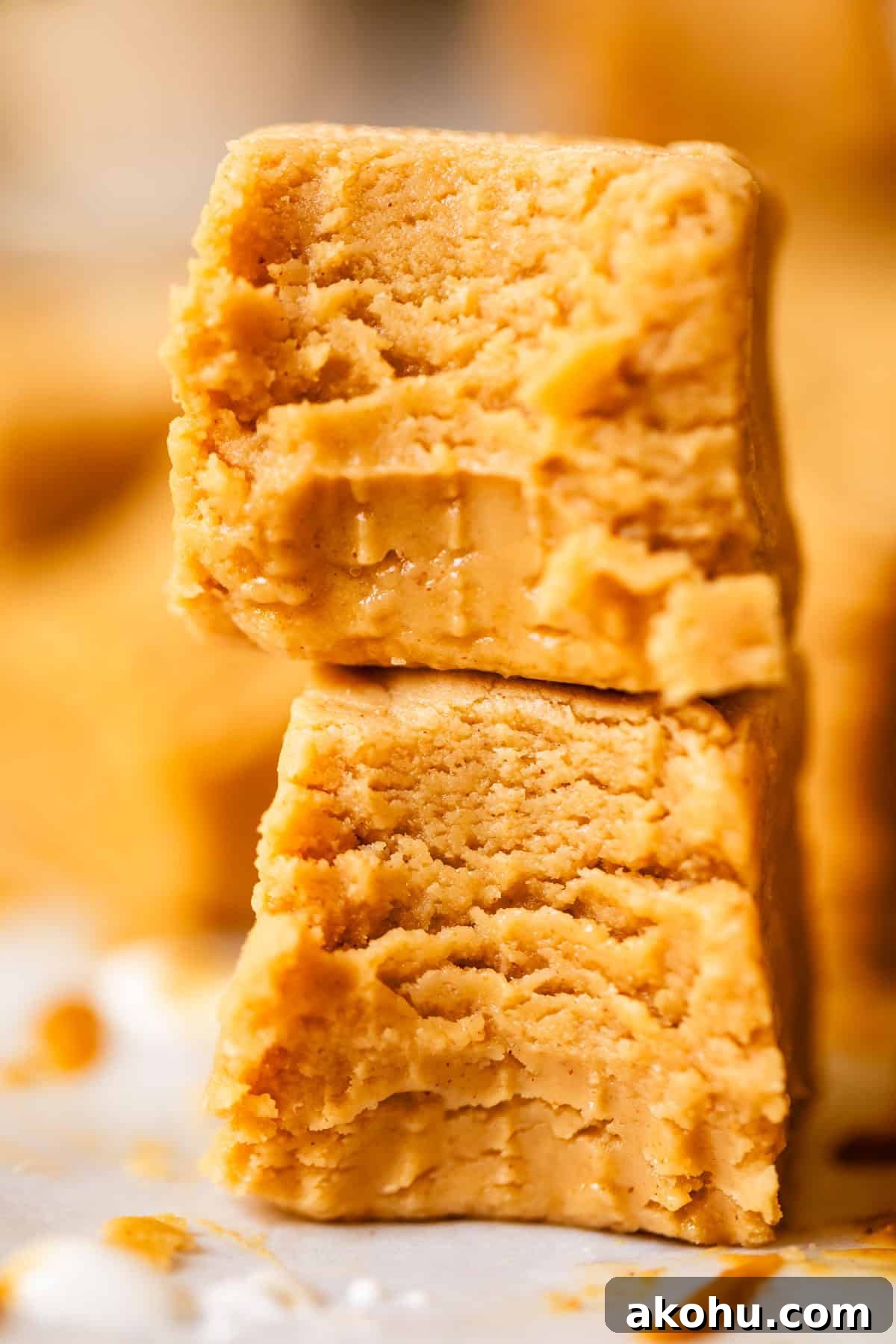 Two thick, creamy, golden-brown squares of peanut butter fudge are stacked on top of each other, showing a soft and smooth texture with some bite marks and crumbly edges.