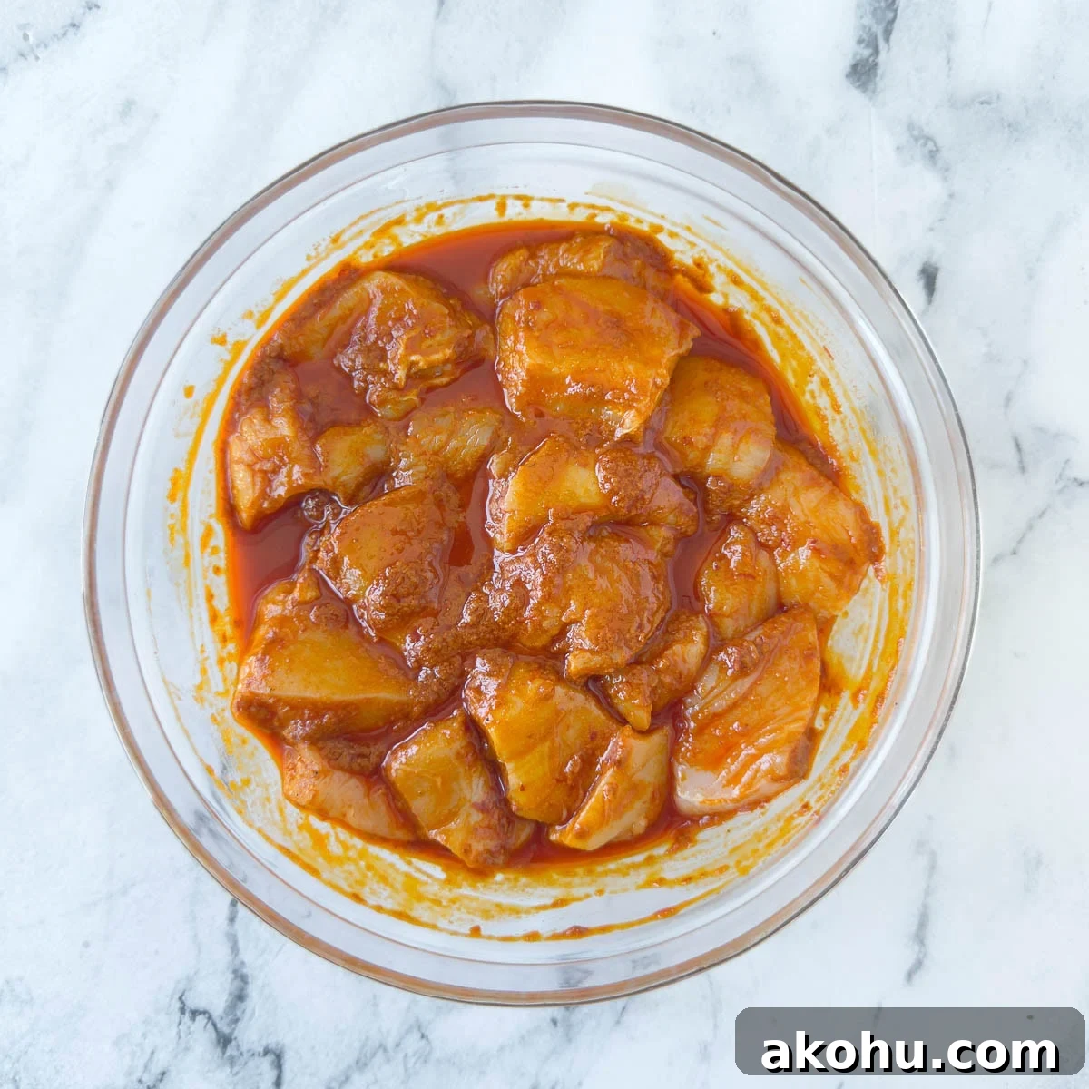 Pieces of chicken breast in a bright orange harissa marinade in a glass bowl.