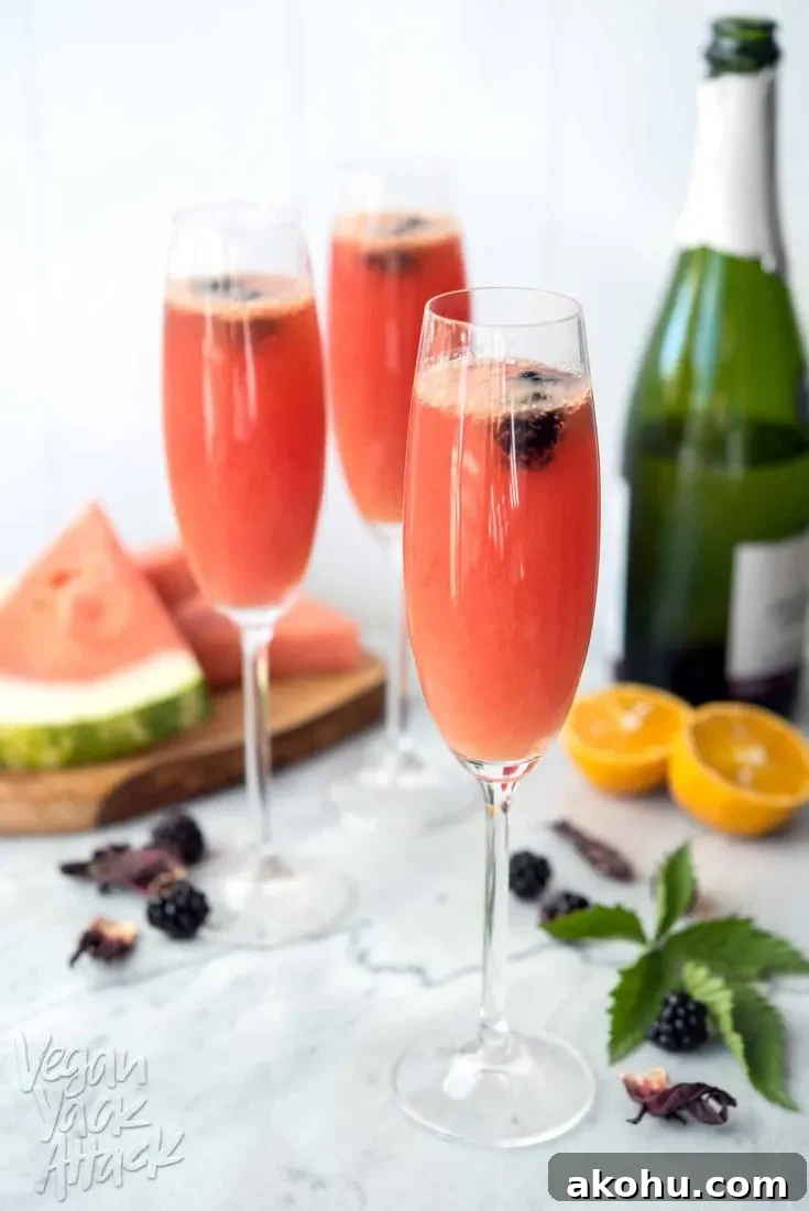 Image of three Champagne glasses filled with watermelon hibiscus mimosas, on a marble table top, surrounded by ingredients.