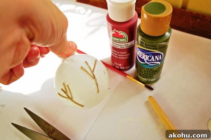 Image shows a hand holding the painted ornament with twine trees, next to two small bottles of paint