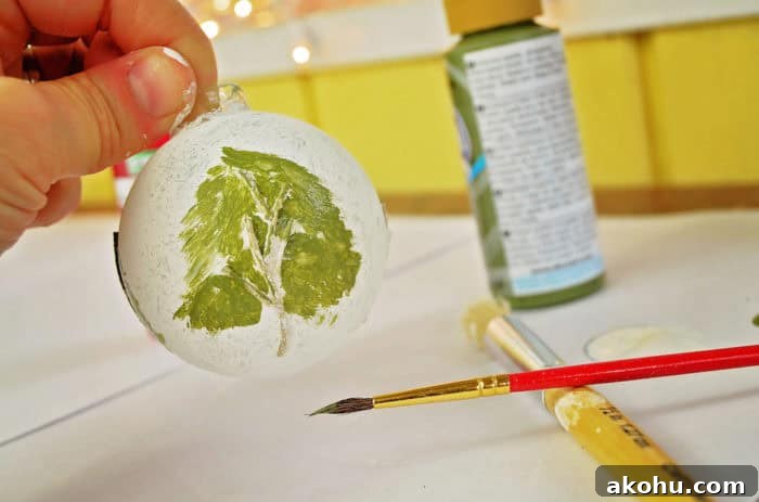 Photo shows a hand holding the painted ornament, while painting the green tree on it