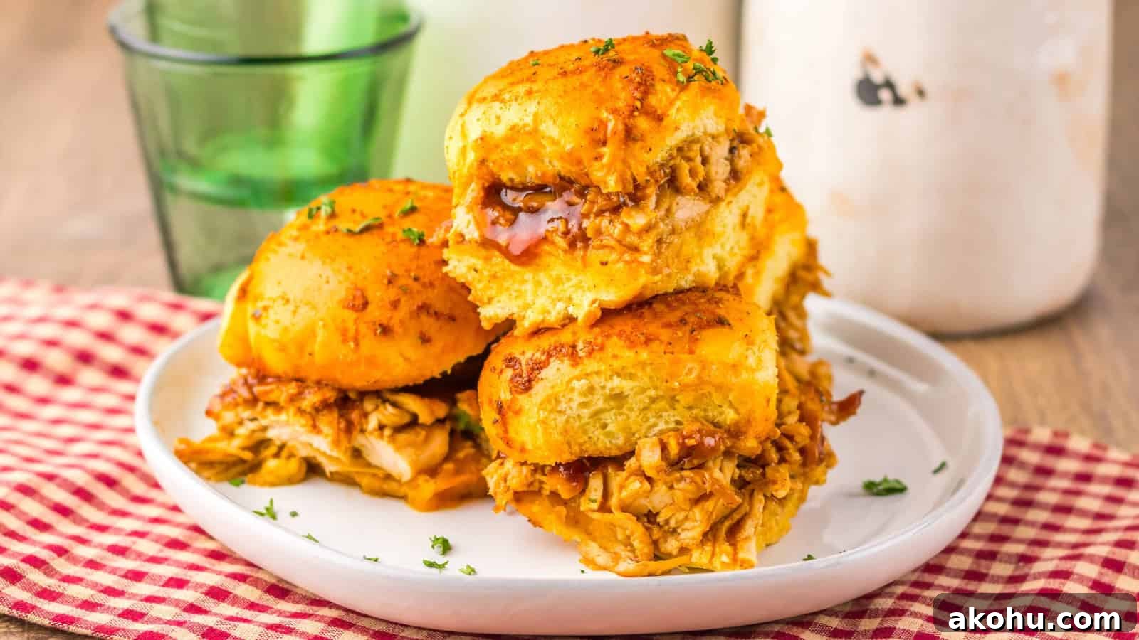 Three BBQ chicken sliders stacked on a white plate, with a red checked cloth and a green glass nearby.