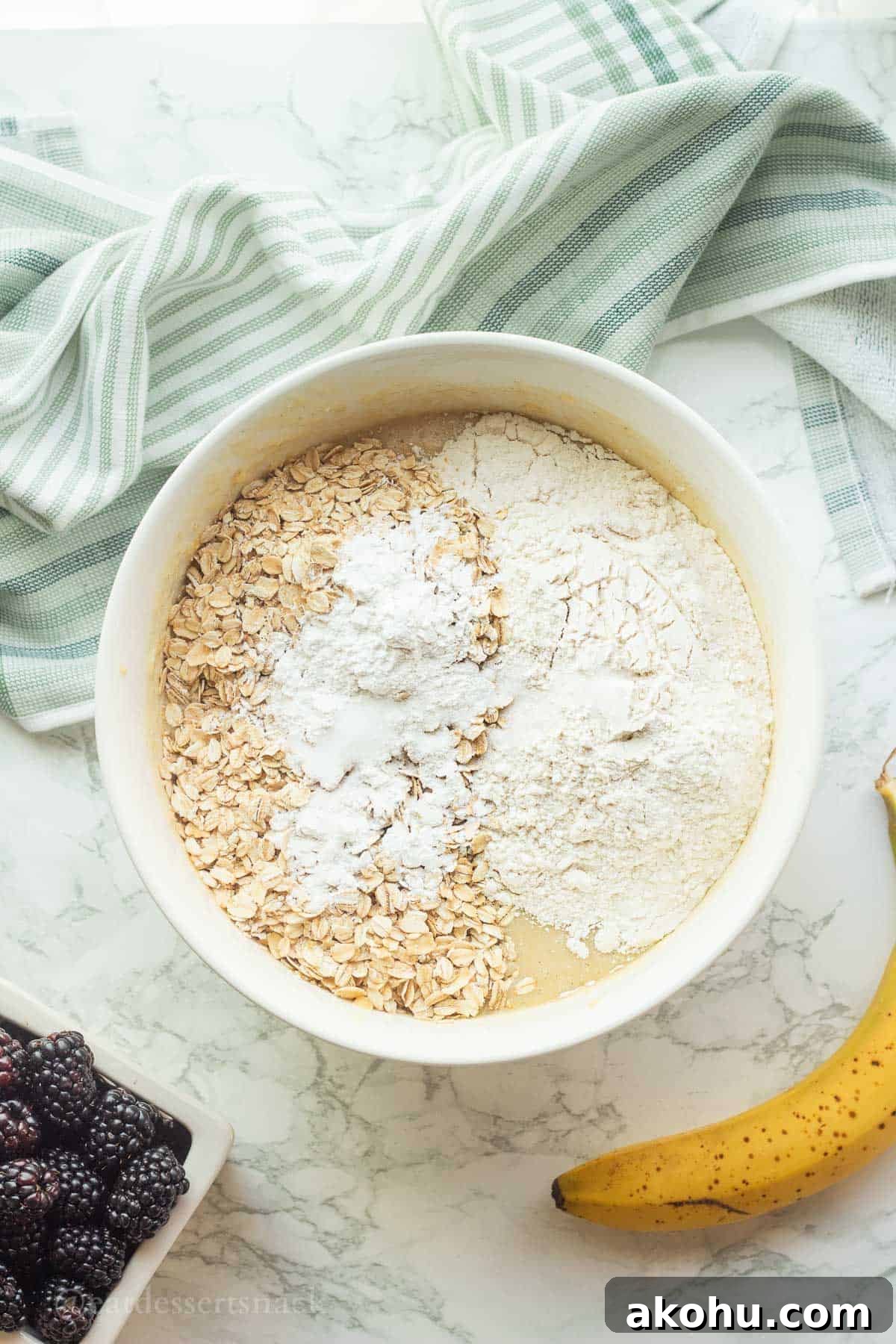 Banana Blackberry Oatmeal Muffins Recipe — Quick Baked Breakfast 7 Flour, oats, and baking powder in white bowl of wet muffin batter.