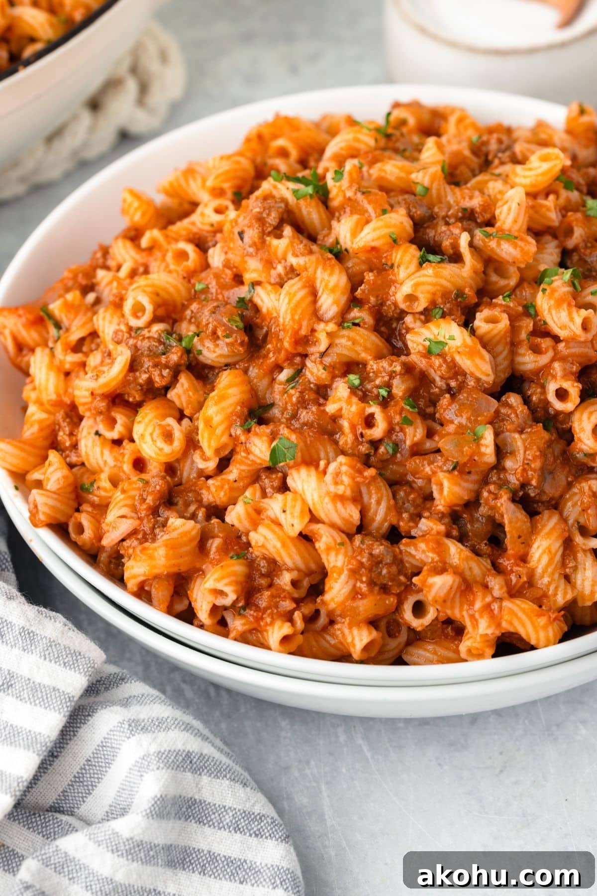 Classic Beefaroni Recipe: Hearty One‑Pot Pasta Dinner 2 A bowl of spiral pasta topped with a rich, savory meat sauce garnished with chopped herbs. The pasta is vibrant orange from the sauce. A striped napkin is visible on the side, and a small bowl of salt is in the background.