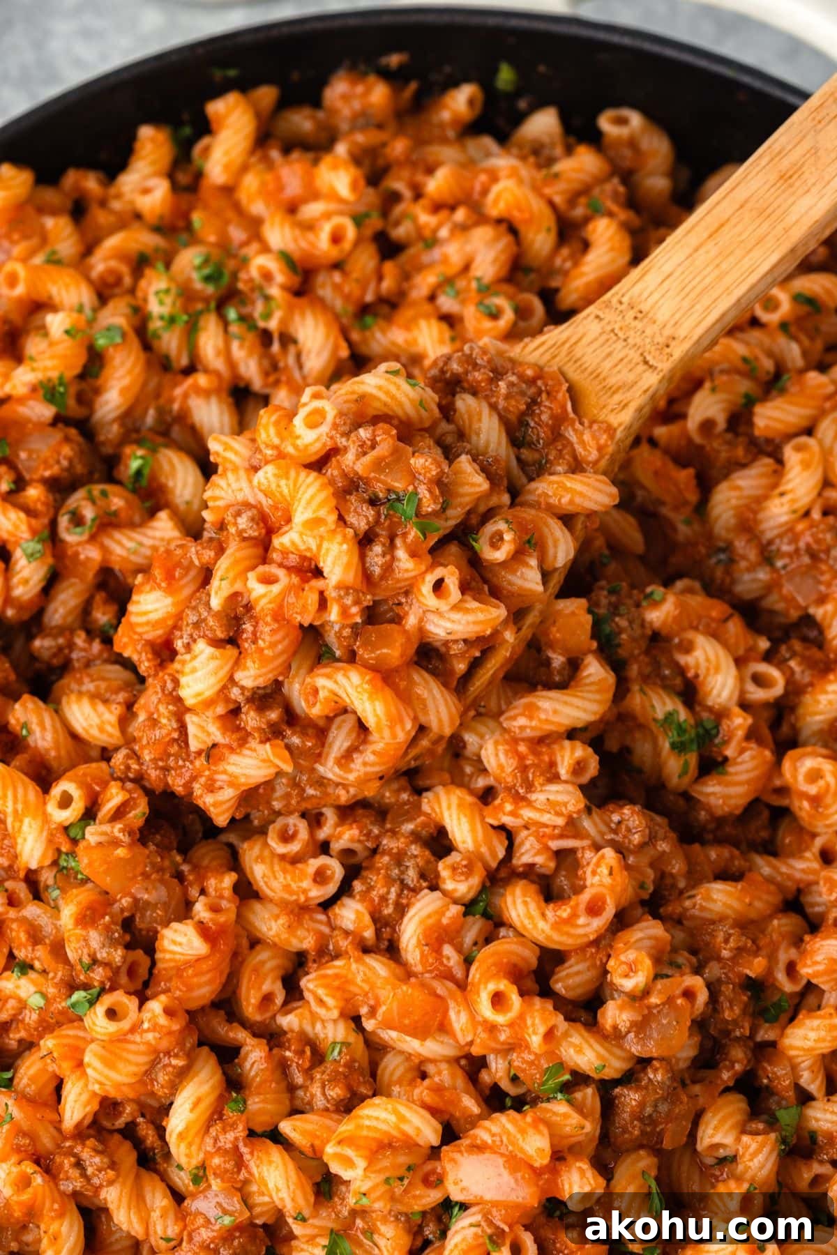 Classic Beefaroni Recipe: Hearty One‑Pot Pasta Dinner 9 A wooden spoon scoops a portion of pasta with a rich, meaty tomato sauce from a black skillet. The dish features curly pasta shapes, garnished with herbs, creating a hearty and appetizing meal.