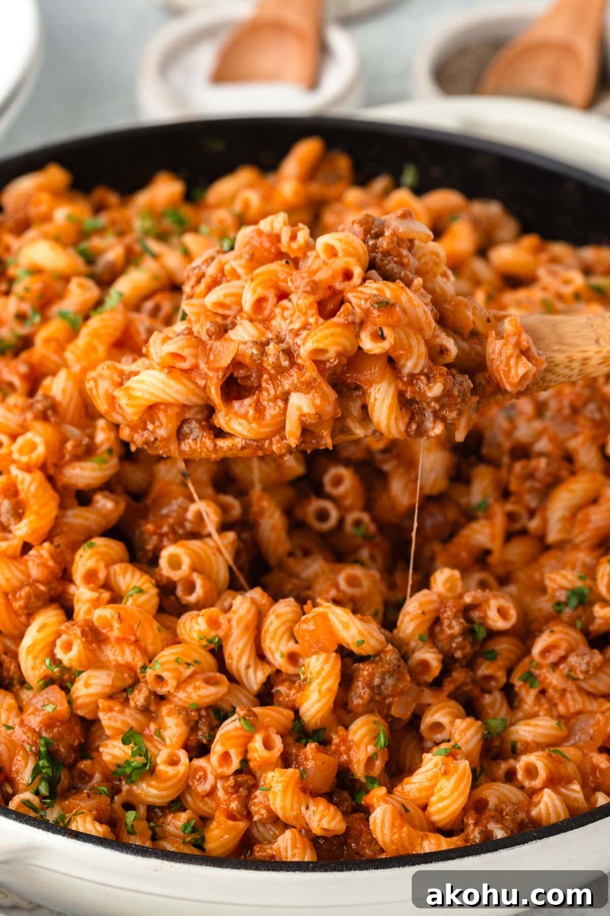 Classic Beefaroni Recipe: Hearty One‑Pot Pasta Dinner 3 Close-up of a skillet filled with cooked cavatappi pasta in a rich, red meat sauce. A wooden spoon lifts a portion, showing noodles and bits of ground meat. Garnished with fresh parsley, the dish looks appetizing and warm.