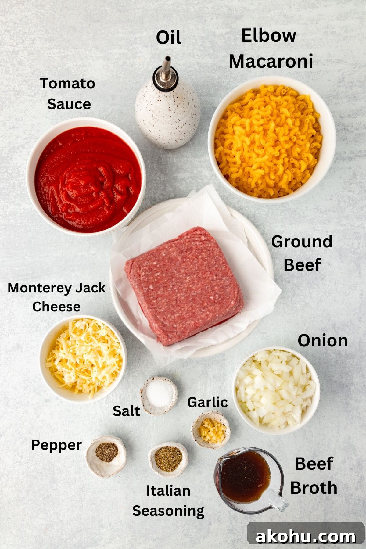 Classic Beefaroni Recipe: Hearty One‑Pot Pasta Dinner 4 Ingredients are arranged on a light surface: bowl of elbow macaroni, ground beef, diced onion, beef broth, tomato sauce, Monterey Jack cheese, and seasonings including garlic, salt, pepper, Italian seasoning, and oil in a bottle.