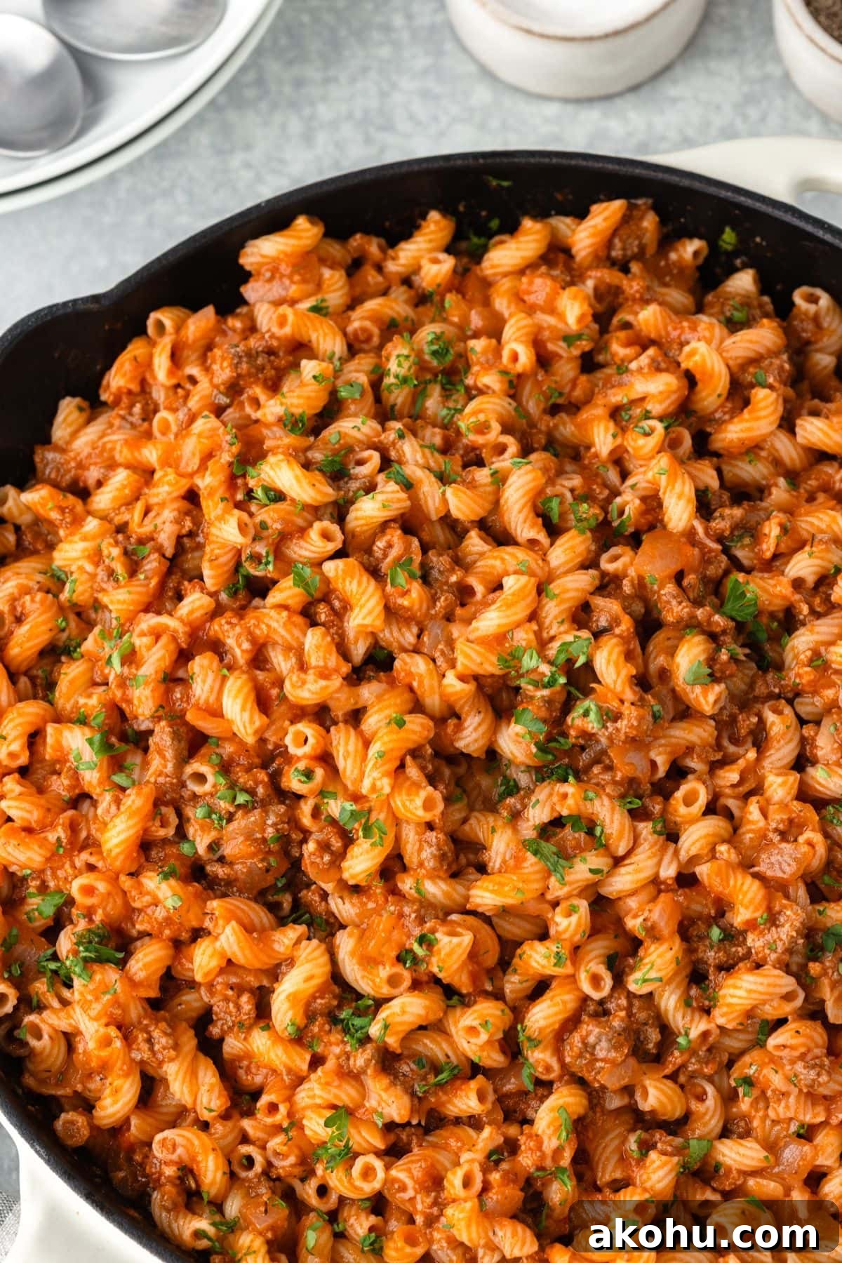 Classic Beefaroni Recipe: Hearty One‑Pot Pasta Dinner 6 A pan filled with cooked pasta mixed with ground meat in a rich red sauce, garnished with chopped parsley. The pasta is spiral-shaped, and the dish appears hearty and flavorful, set on a light-colored surface.