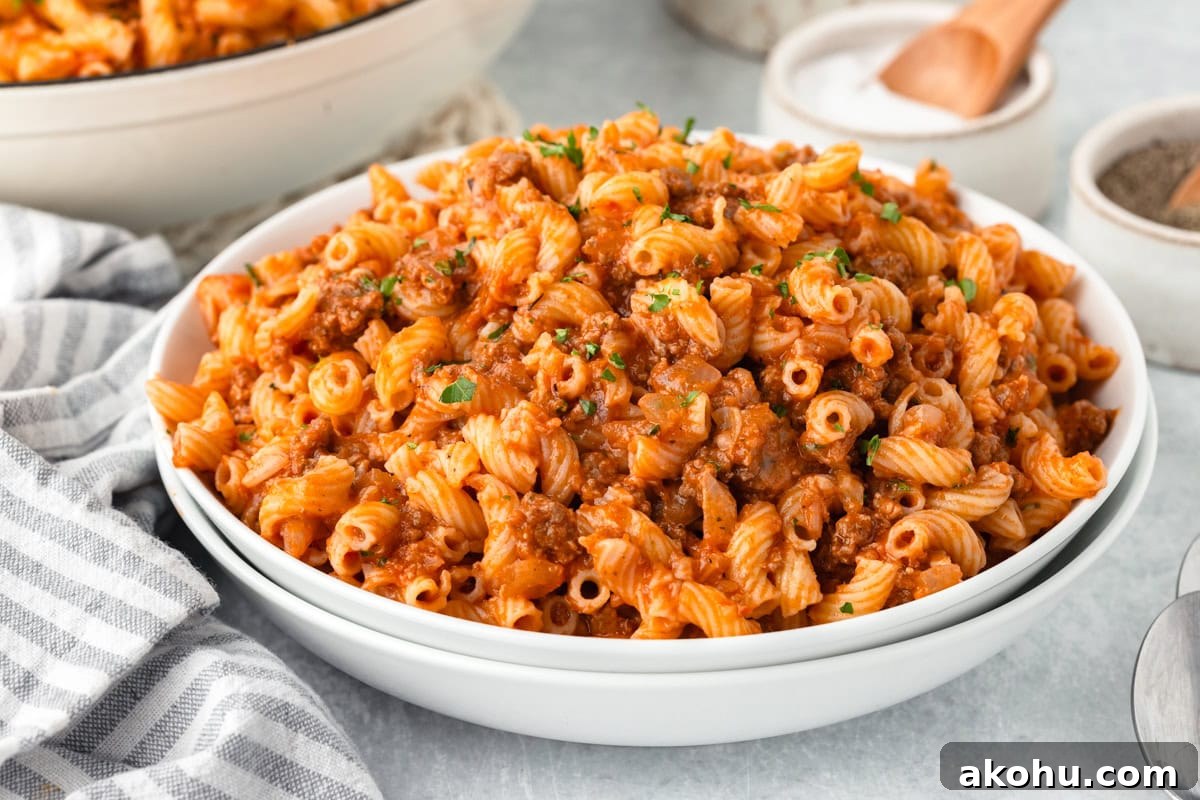 Classic Beefaroni Recipe: Hearty One‑Pot Pasta Dinner 7 A white bowl filled with cooked pasta mixed with a rich tomato and meat sauce, garnished with chopped parsley. A striped napkin is on the side, and small bowls of salt and pepper are nearby on a light-colored surface.