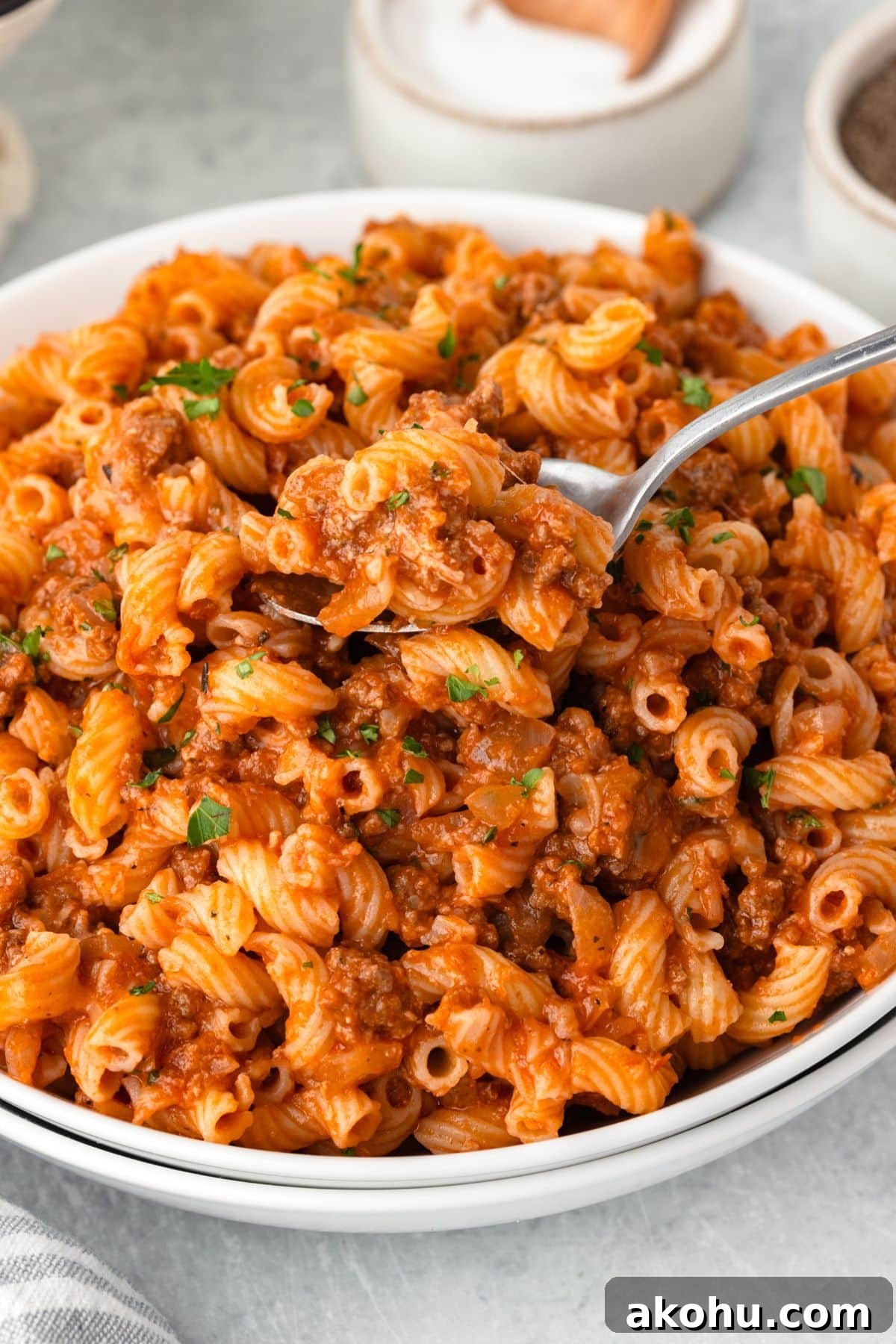 Classic Beefaroni Recipe: Hearty One‑Pot Pasta Dinner 8 A bowl of rotini pasta topped with a rich, tomato-based meat sauce. A spoon lifts a portion of the pasta, which is garnished with chopped parsley. Salt and pepper shakers are visible in the background on a light-colored surface.