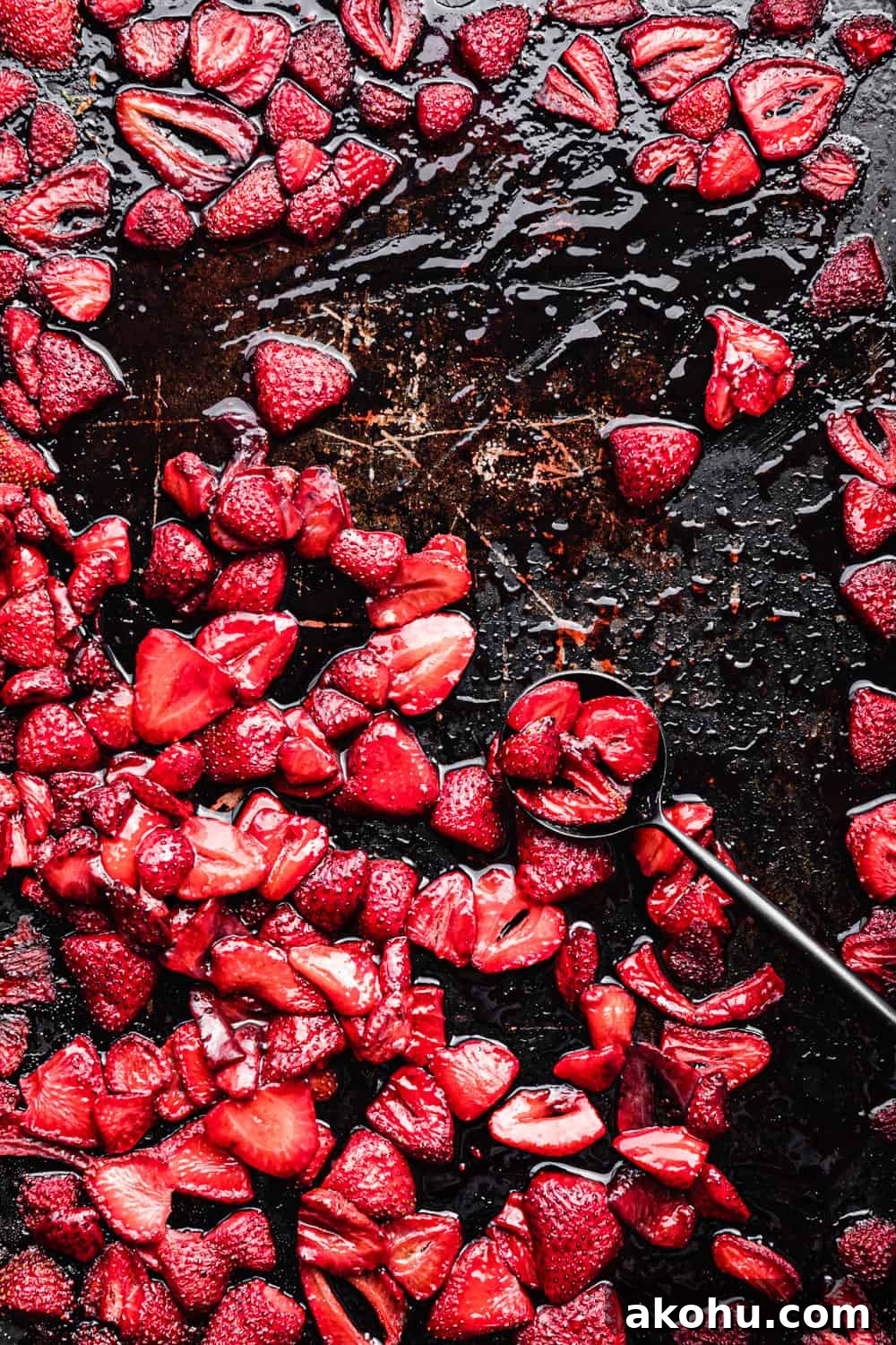 Overhead shot of jammy roasted strawberries in their syrup on a dark sheet pan with a black spoon, post-oven, on a dark background.