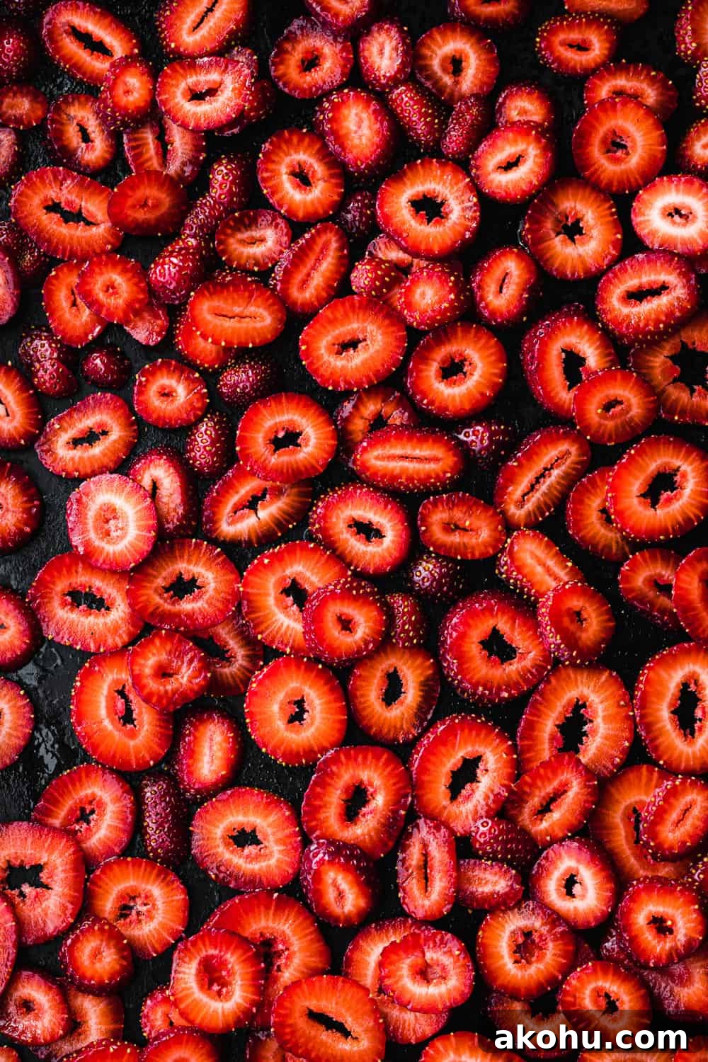 Overhead shot of sliced strawberries on a dark sheet pan before roasting, arranged in a single layer showing their vibrant red flesh and seeds.