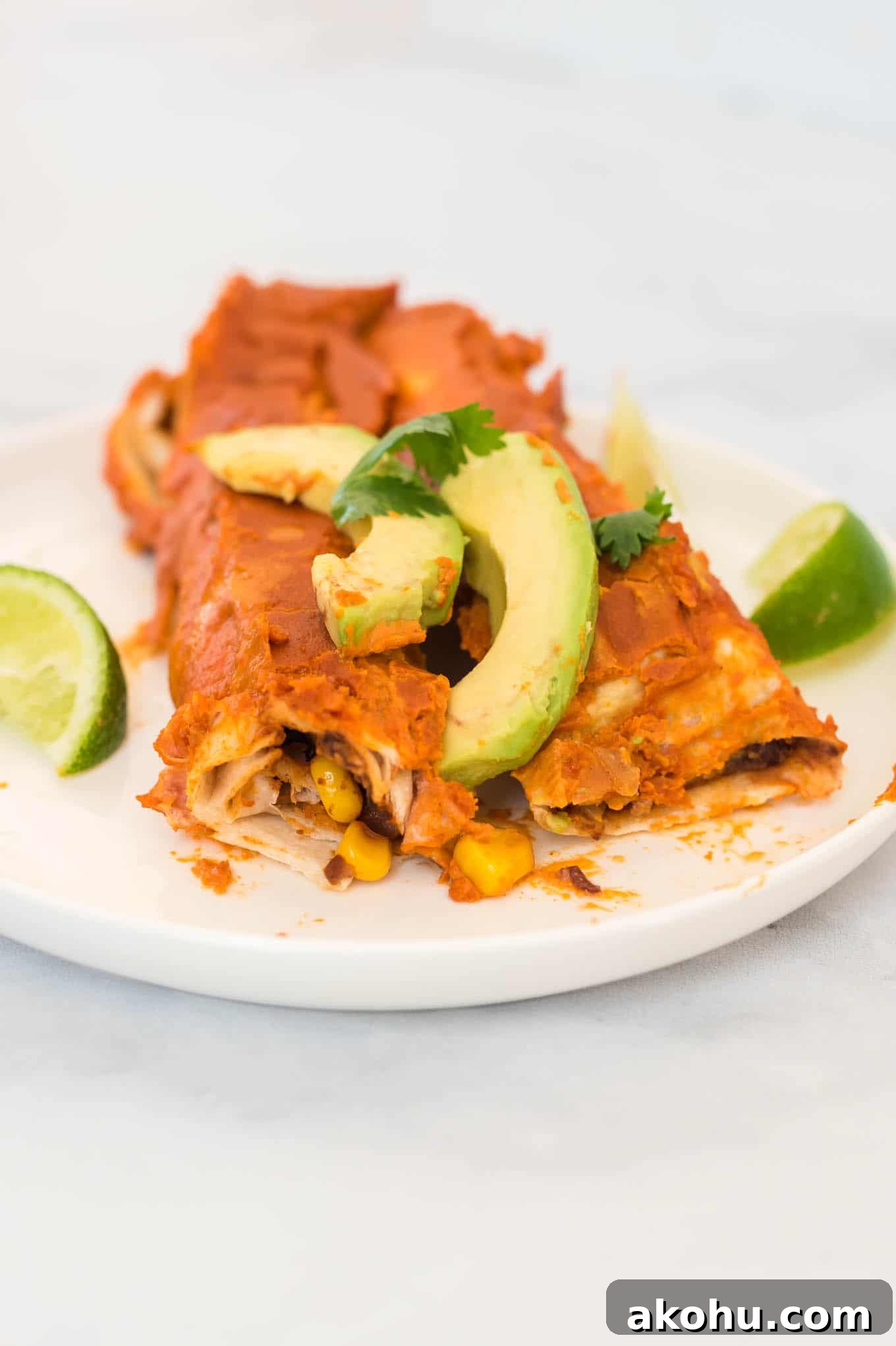two enchiladas on a white plate topped with avocado and cilantro, with limes on the side