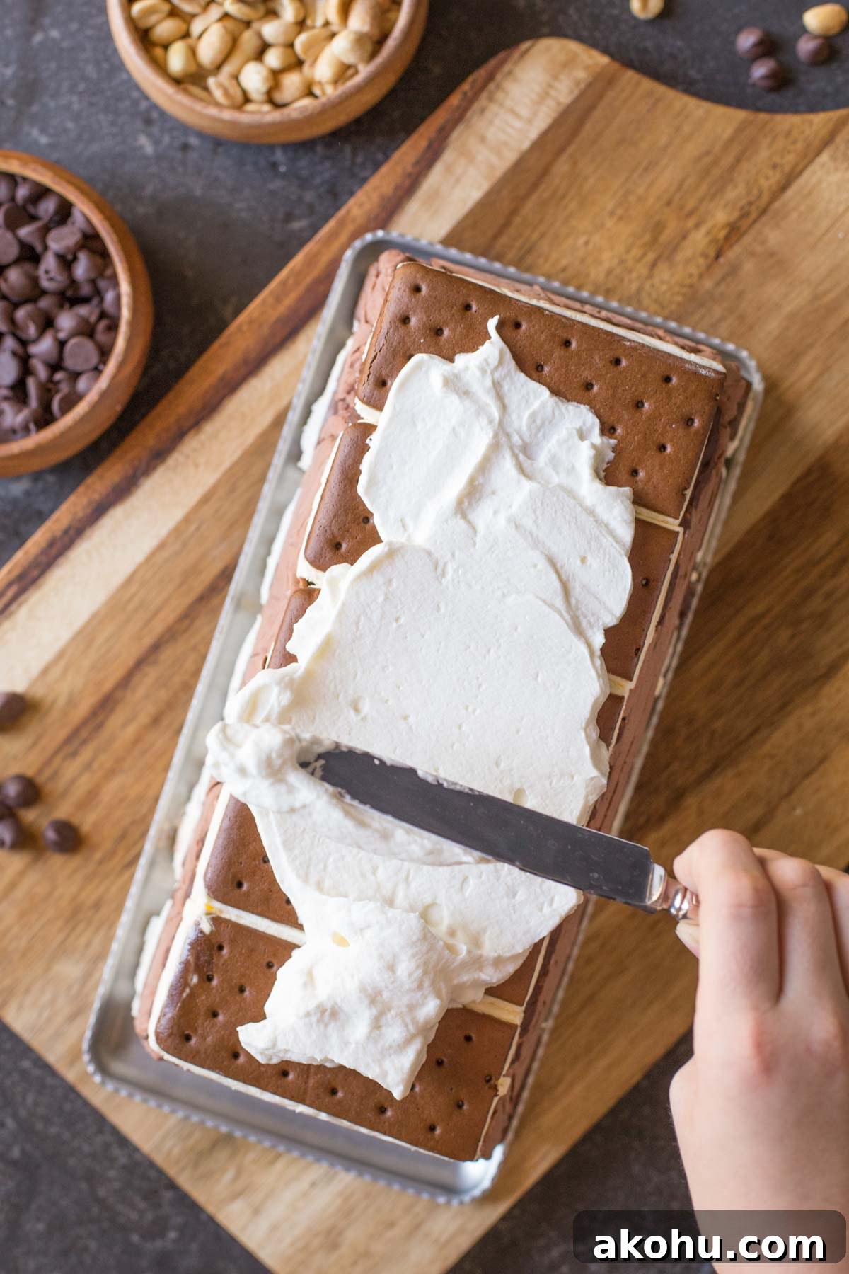 Chocolate Peanut Butter Ice Cream Slice Cake Recipe 5 Whipped cream being spread onto the Chocolate Peanut Butter Ice Cream Slice Cake, that is sitting on a platter on top of cutting board, with a bowl of chocolate chips and a bowl of peanuts next to it.