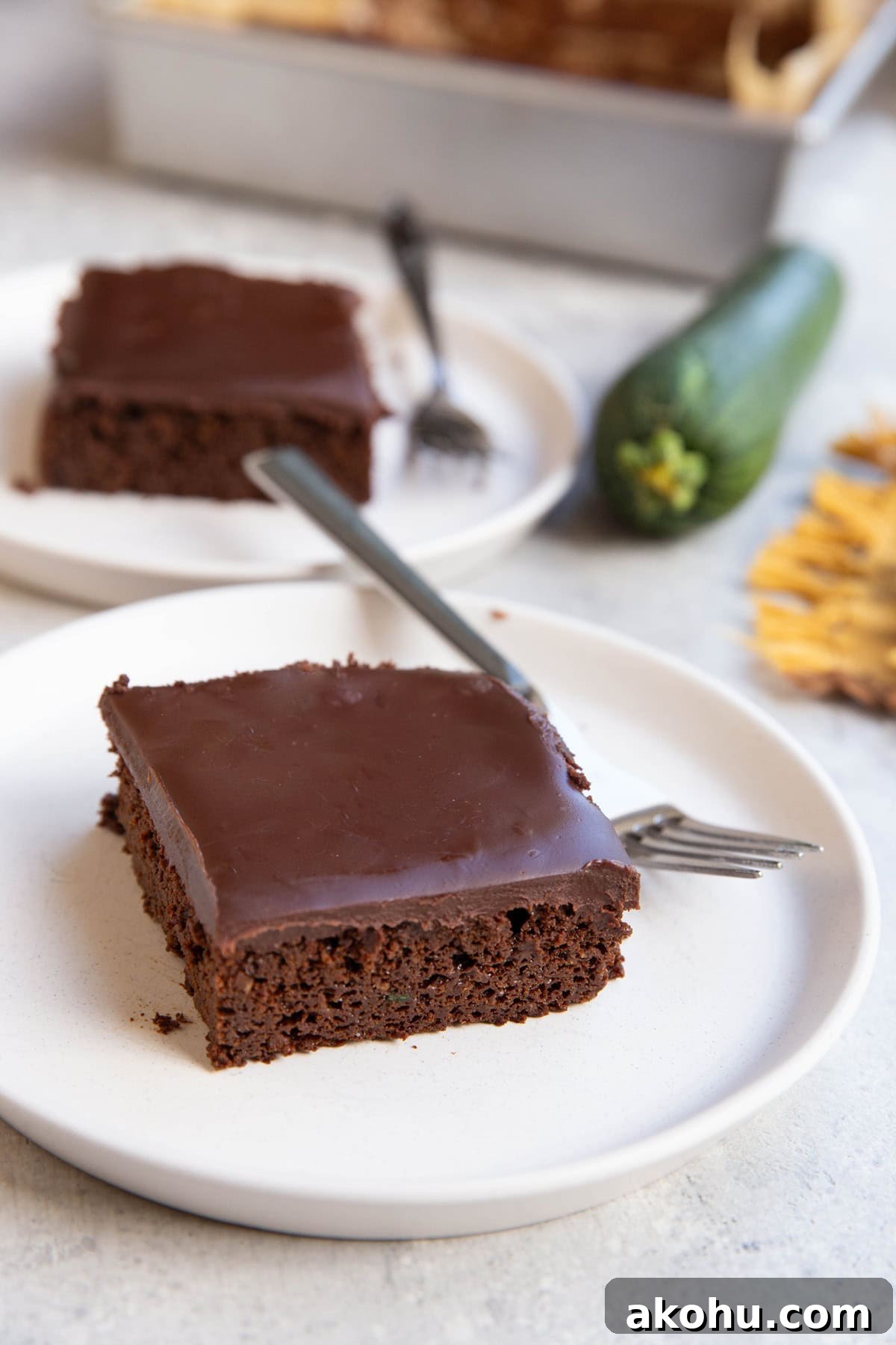 Two slices of chocolate zucchini cake covered frosted with chocolate frosting and a fresh zucchini in the background.