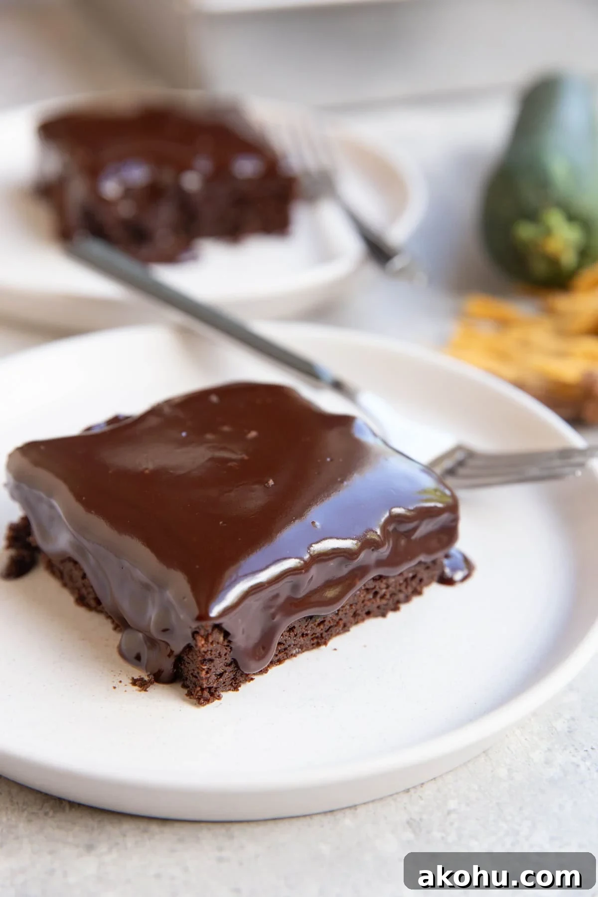 Two slices of zucchini chocolate cake with chocolate ganache melting down the top and sides on white plates and forks, ready to serve. A zucchini squash and a golden napkin in the background.