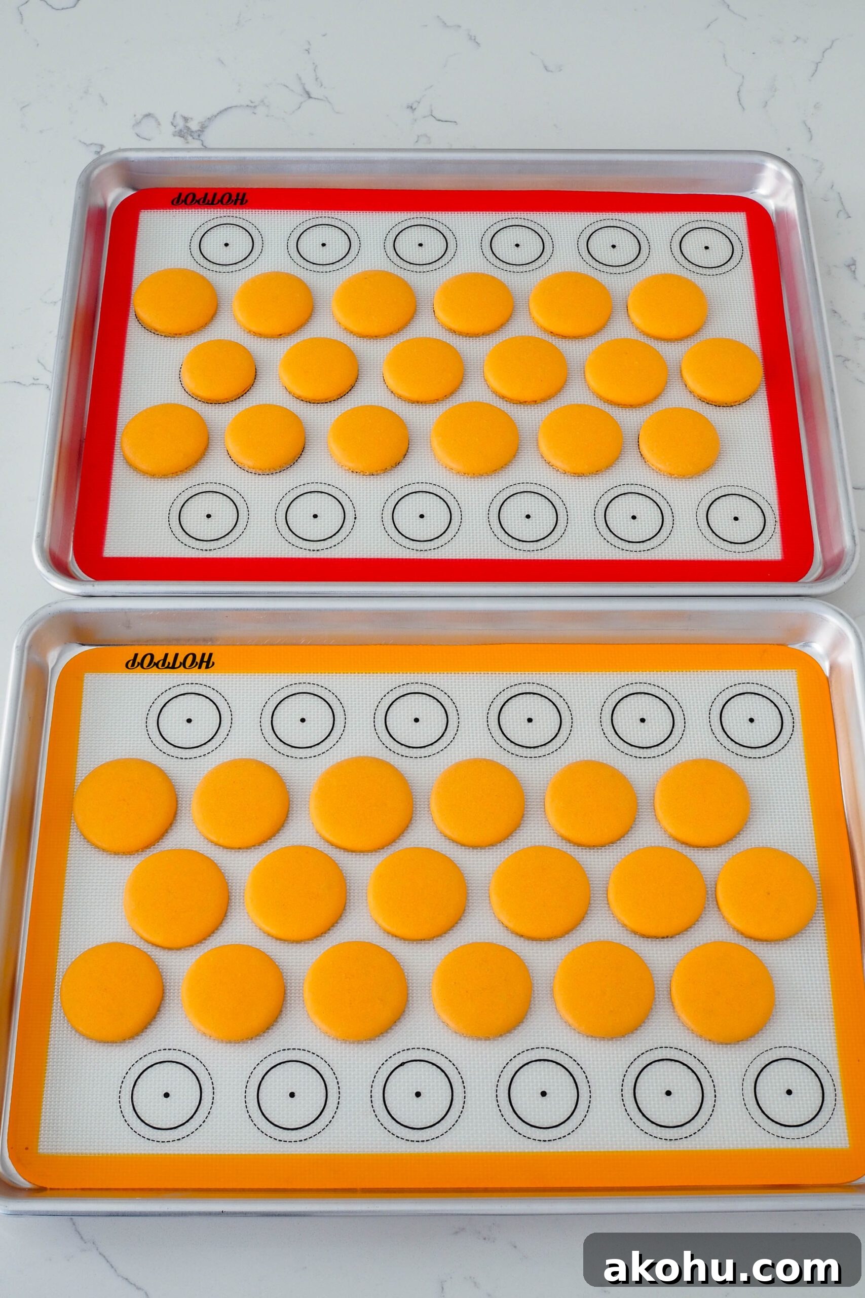 Two trays of baked orange macaron shells on silicone mats and aluminum pans.