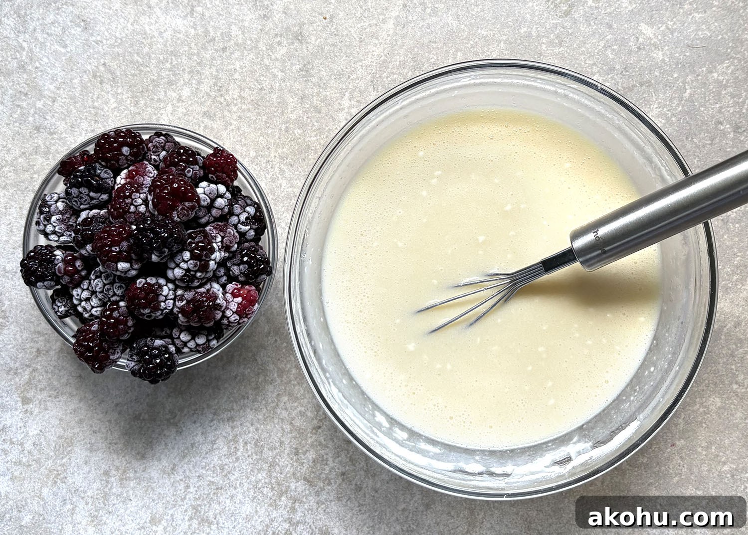 Blackberry cobbler batter mixed in a glass bowl with a whisk and frozen blackberries beside it.