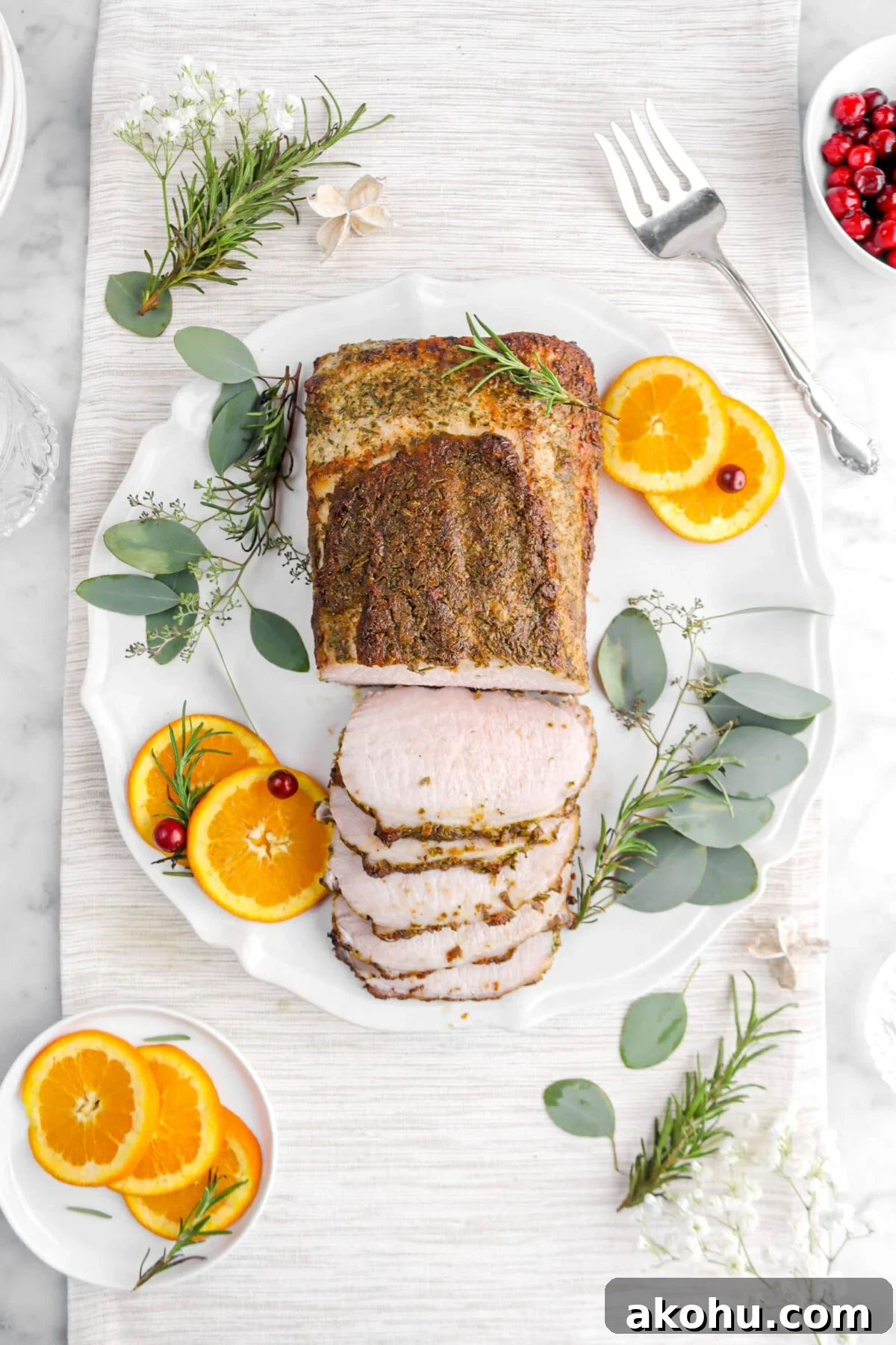 overhead shot of pork tenderloin with five slices laying in front on round platter with orange slices, rosemary sprigs, eucalyptus leaves, and cranberries around.