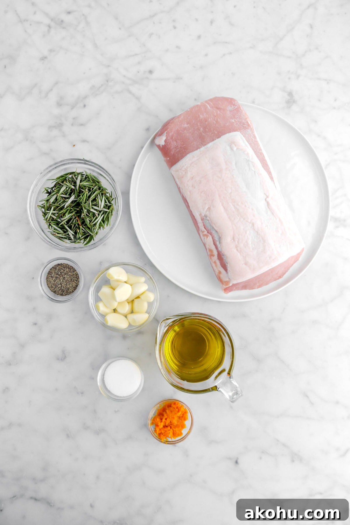 raw pork tenderloin, rosemary leaves, ground black pepper, garlic cloves, olive oil, salt, and orange zest on marble surface.