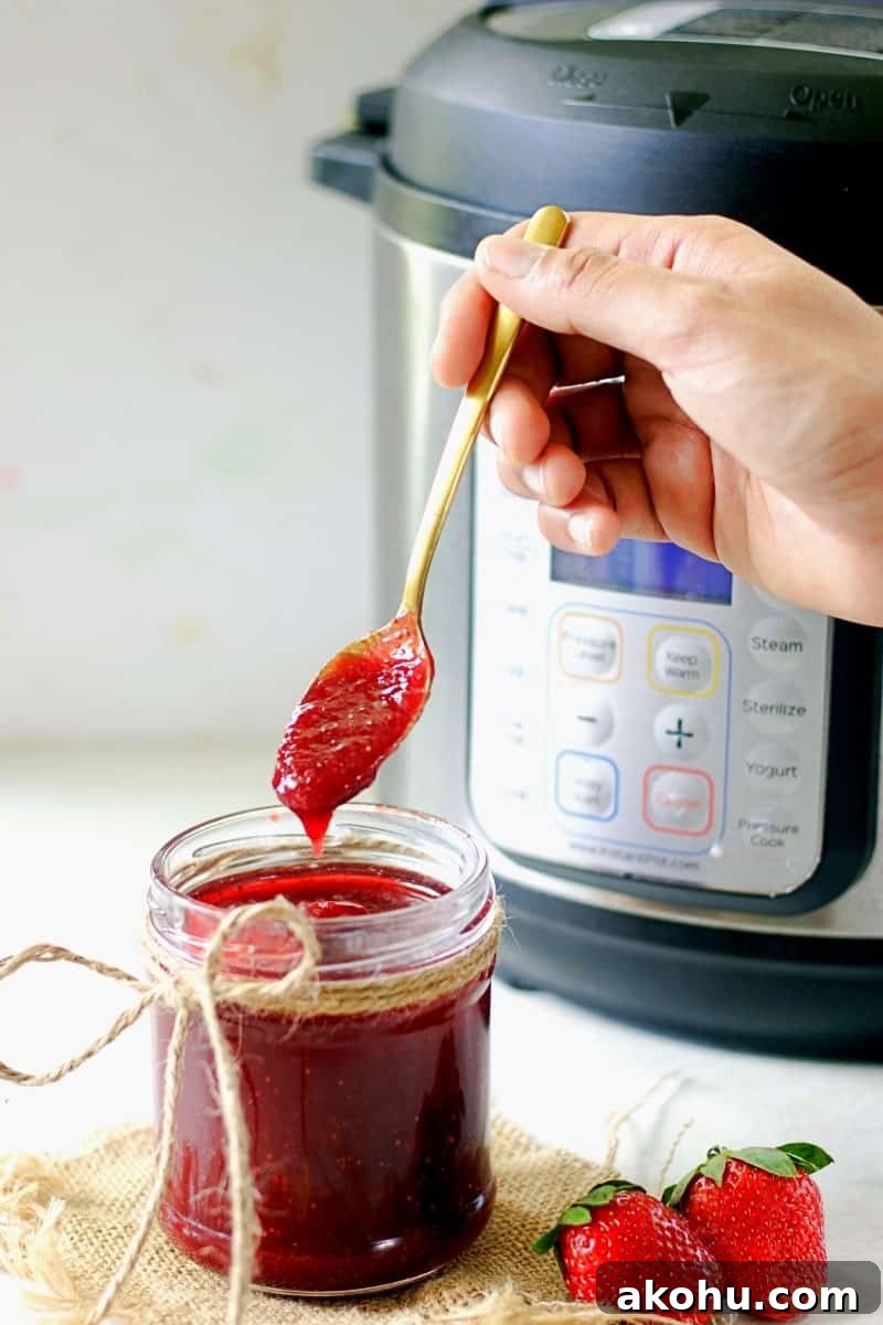 Holiday Instant Pot Strawberry Jam Recipe 9 my fingers are holding a spoon with strawberry jam made in the instant pot which is dripping into the mason jar showing the texture with an instant pot int he background.